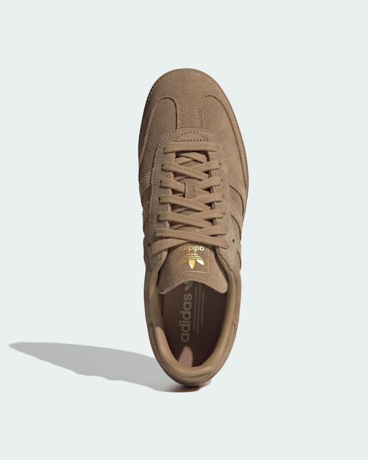 Ανοιχτό καφέ σουέτ Adidas πάνινο παπούτσι με ταιριαστά κορδόνια, λογότυπο χρυσού adidas στη γλώσσα και ένα κολάρο με γεμισμένη. Το παπούτσι έχει ένα κλασικό σχέδιο με ορατή ραφή και μια σκιά που ρίχνει στα αριστερά.